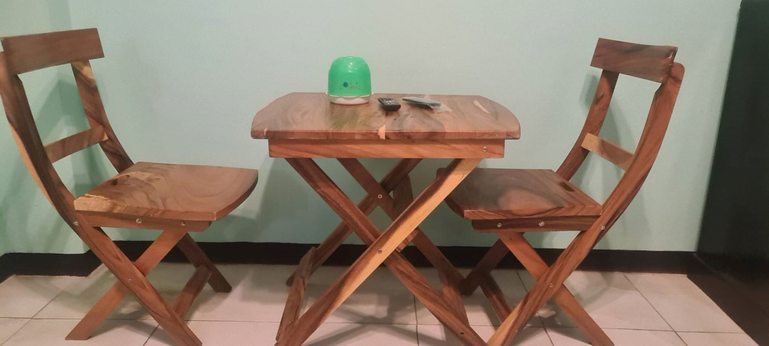 Una pequeña mesa de madera con dos sillas, una tetera verde y un control remoto encima.