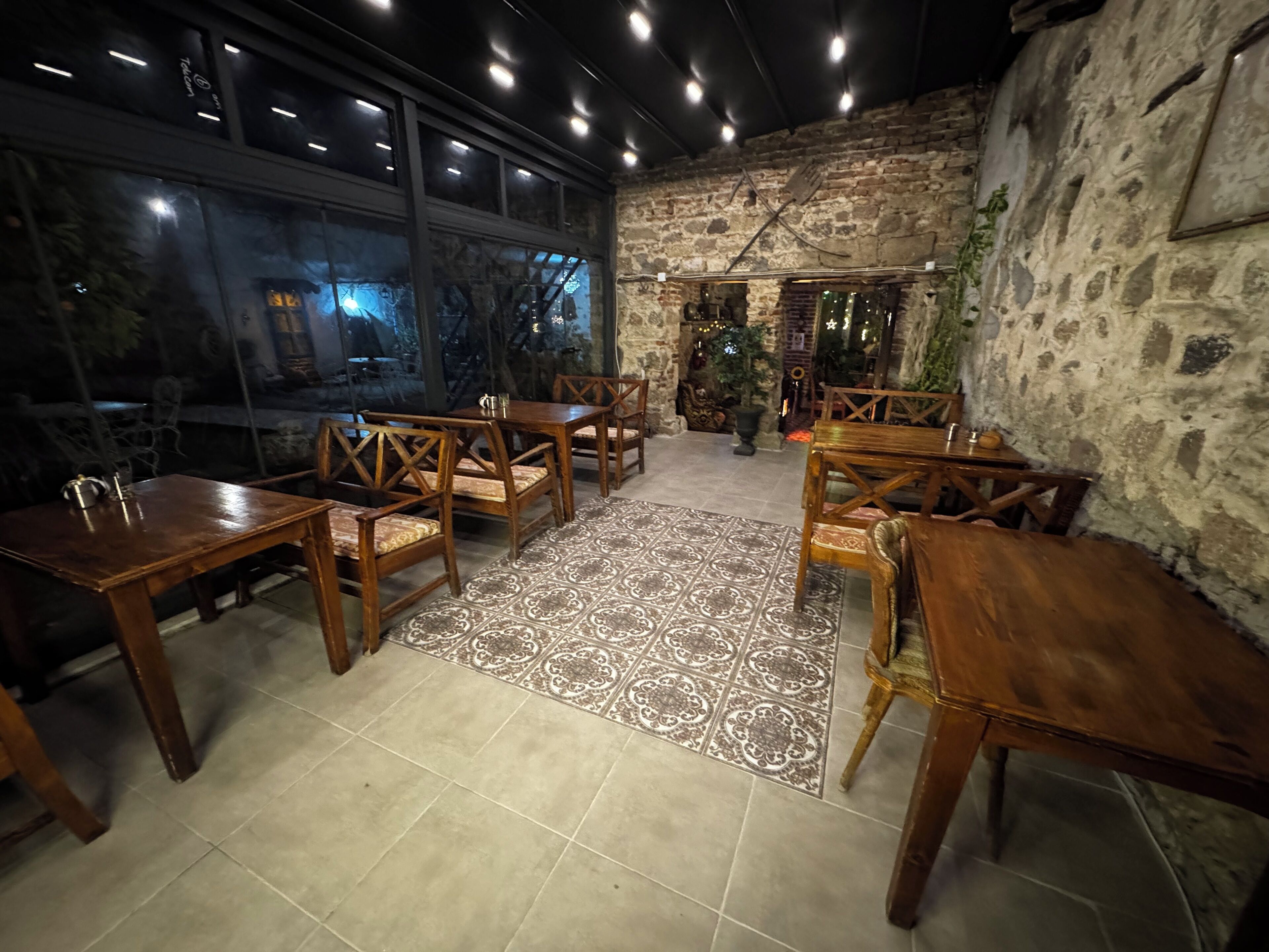 An indoor dining area with wooden tables and chairs, a patterned rug, and a stone wall.