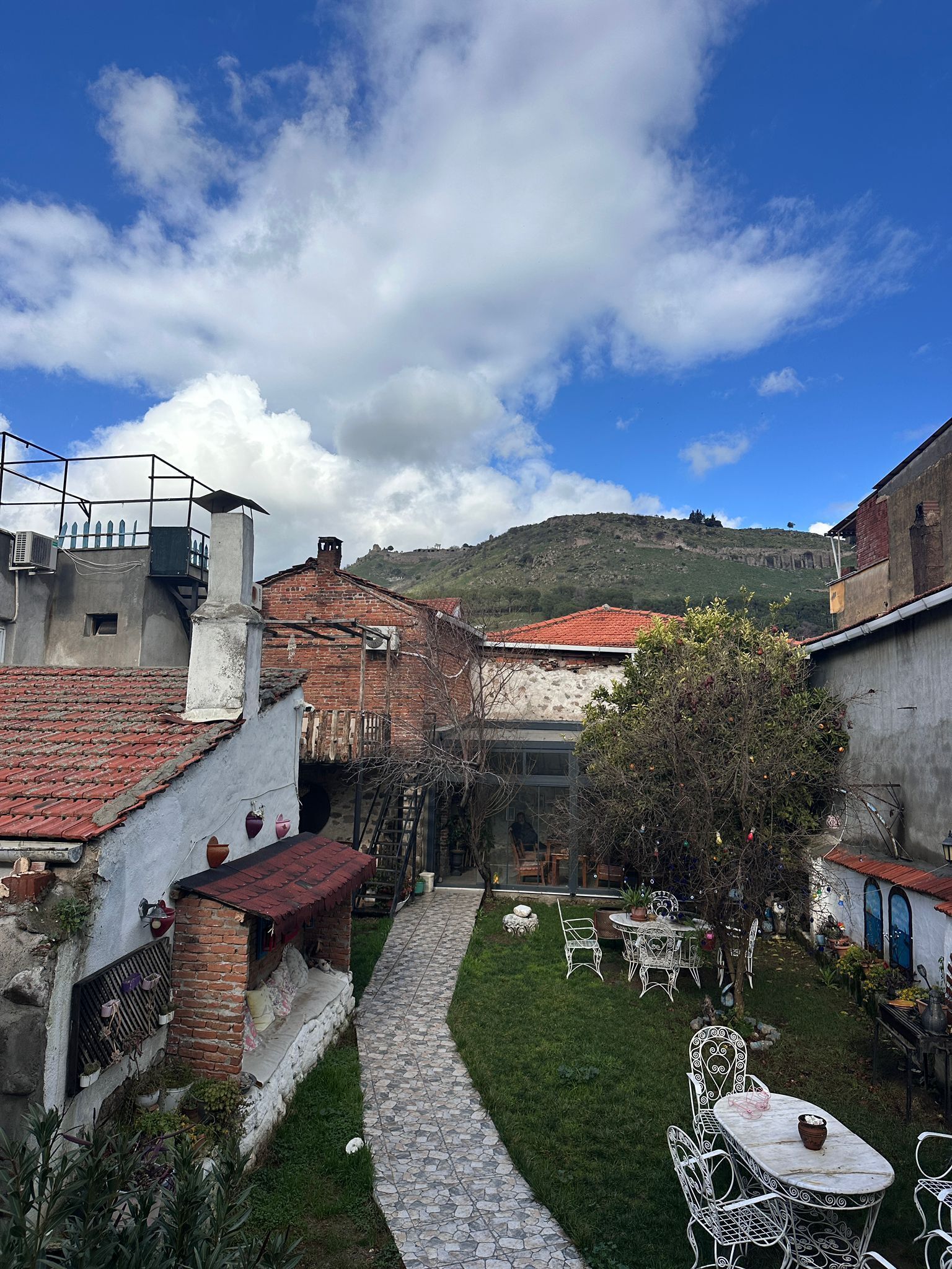 A courtyard with a stone pathway, outdoor seating, and a view of a hill with ruins in the background.