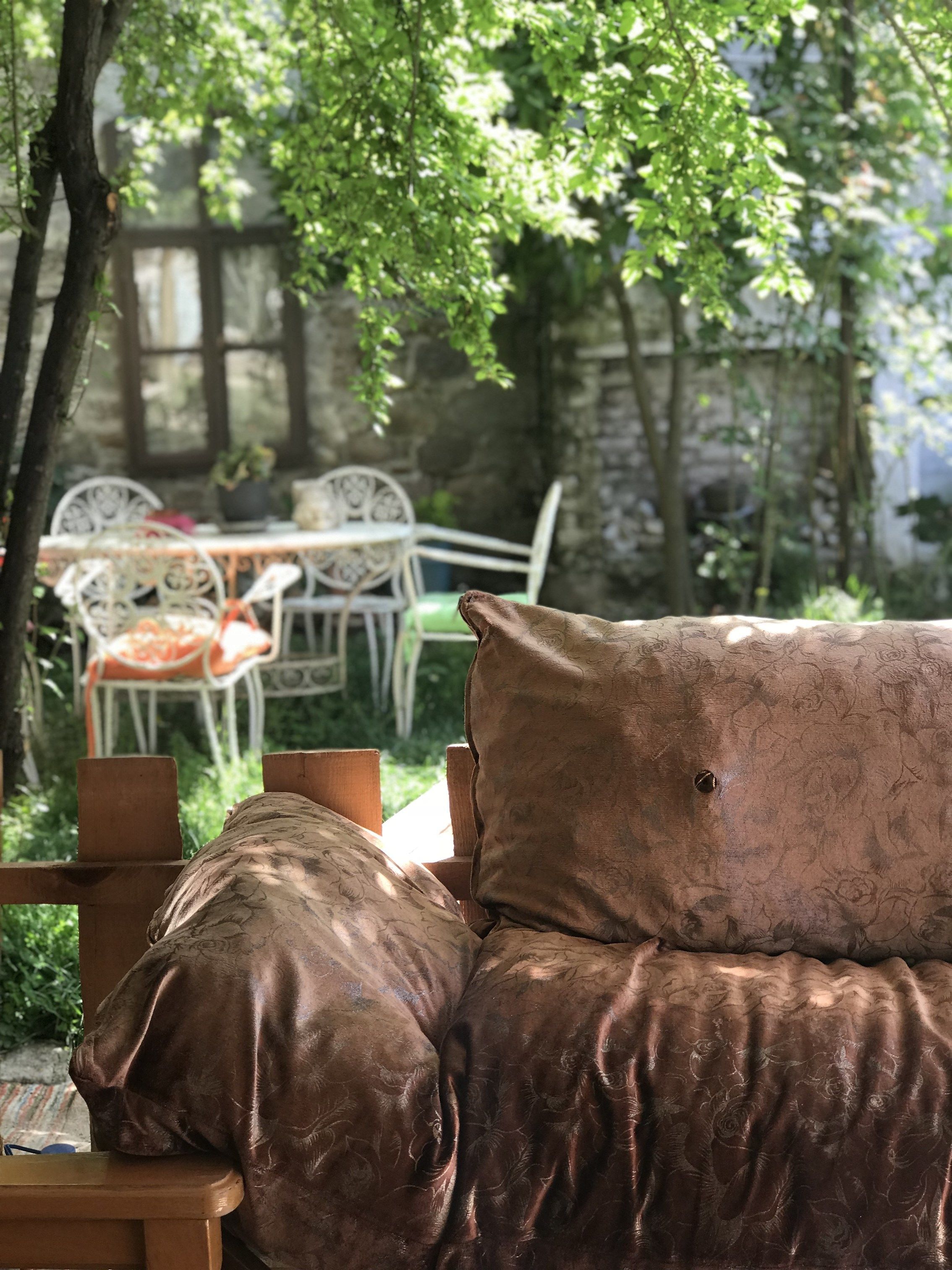 A cozy outdoor seating area with a cushioned bench and a table, surrounded by greenery and stone walls.