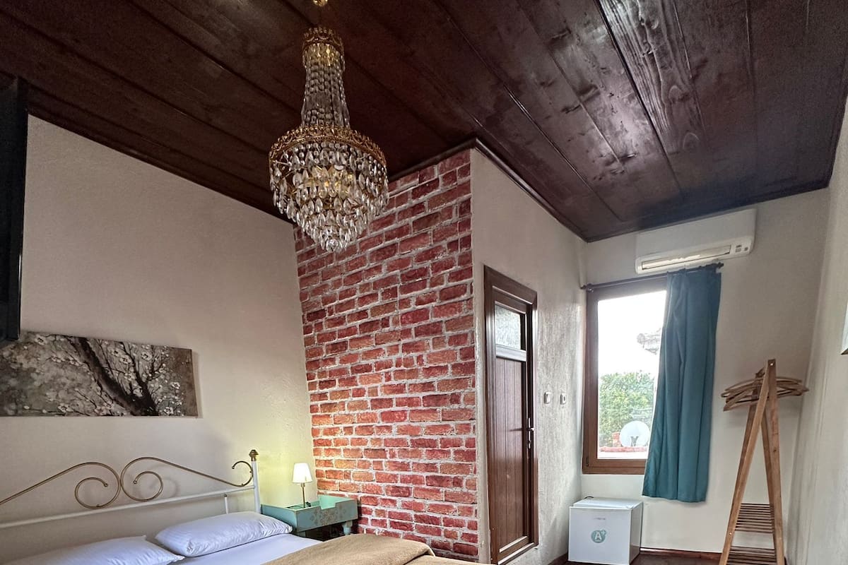 Ein Schlafzimmer mit Holzboden, einer Ziegelwand, einem Bett mit beiger Decke, einer Kronleuchter und einem Fenster mit Vorhängen, sowie einer Holzleiter.