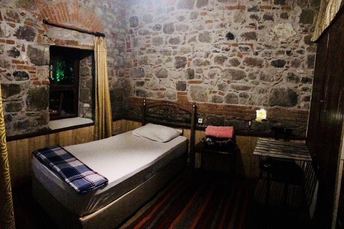 Un lit simple dans une pièce aux murs de pierre, une fenêtre avec des rideaux, une petite table avec une lampe et un plancher en bois.