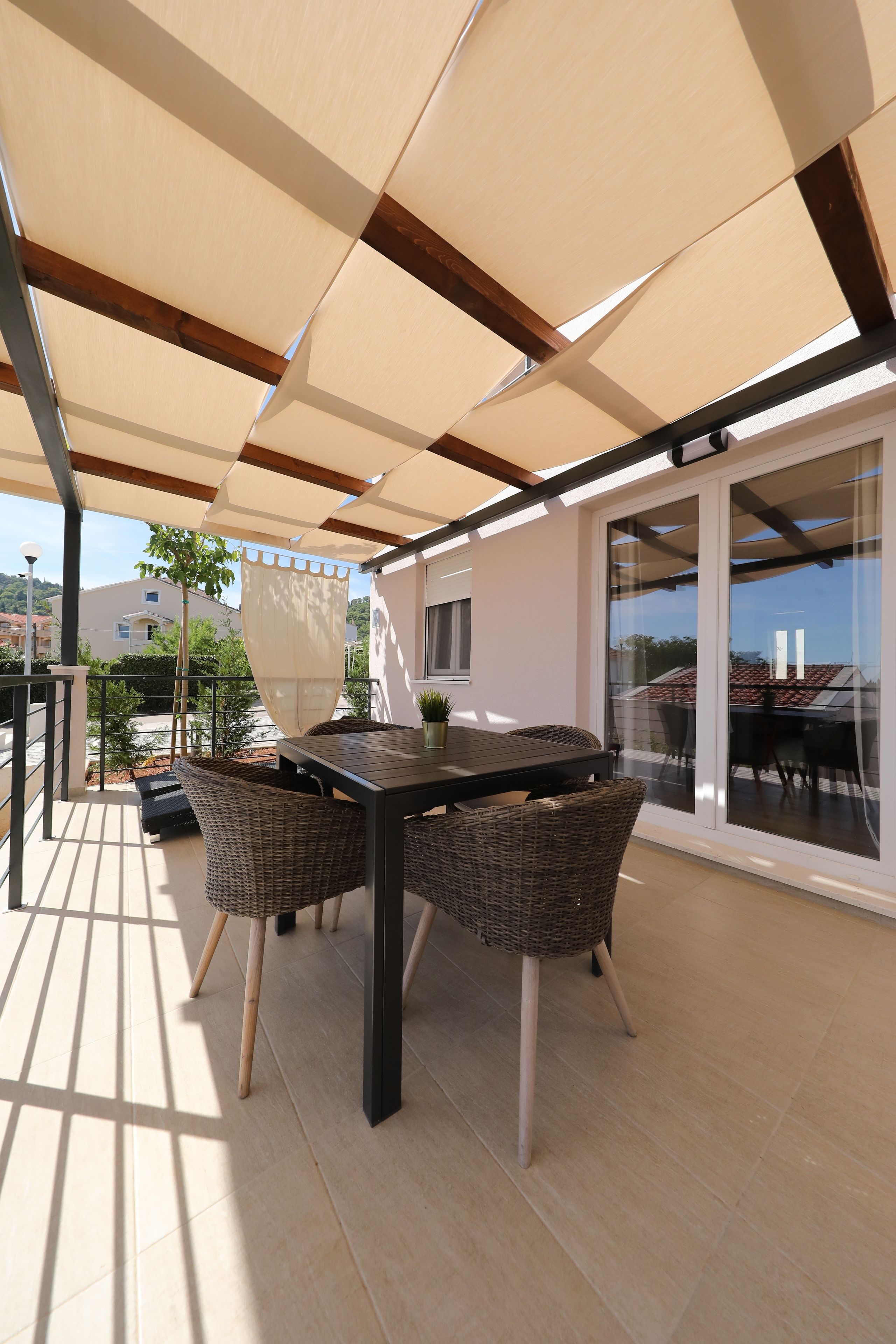 A covered outdoor patio with a table and wicker chairs, a potted plant, and a view of a neighboring building.