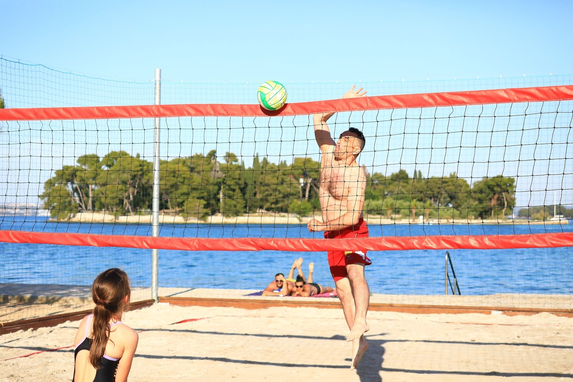 Un match de volleyball de plage avec un filet et des joueurs, une plage de sable, et une étendue d'eau en arrière-plan.