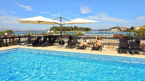 Une piscine avec des chaises longues et des parasols, une vue sur la mer et, au loin, une côte bordée de bâtiments.