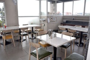A dining area with tables and chairs, a coffee pot, and a window with a view of buildings. - FabHotel Brook Elan Viman Nagar (Pune)