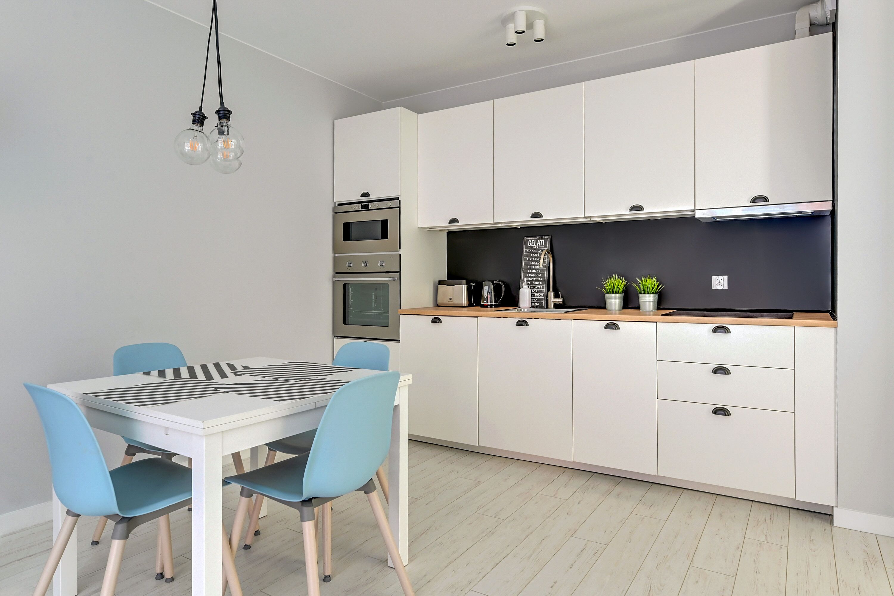 A modern kitchen with white cabinets, a black countertop, and a built-in oven. There is a dining table with blue chairs and a hanging light fixture.