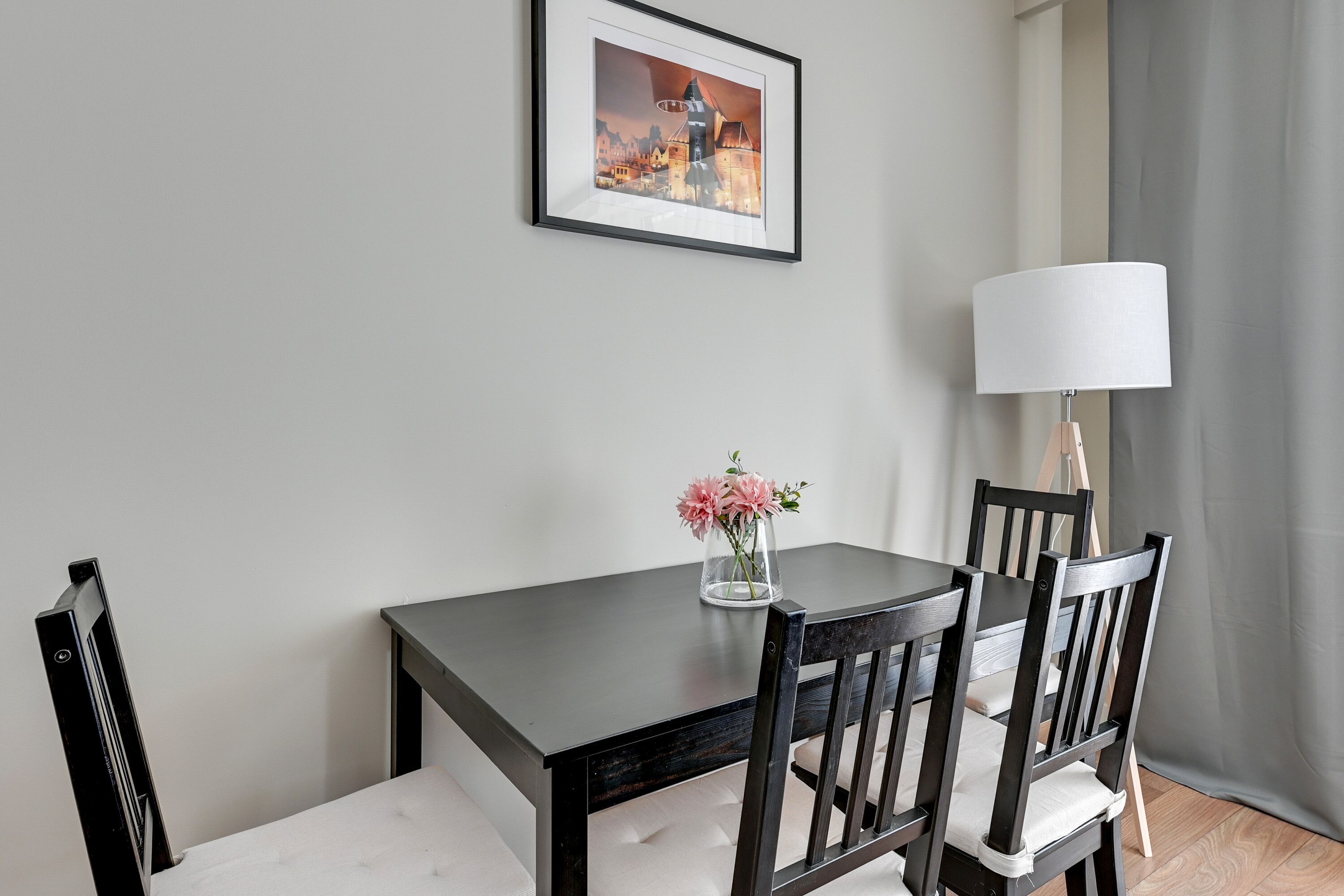 Uma área de refeições com uma mesa de madeira escura, duas cadeiras, um vaso com flores, um quadro na parede e um abajur.