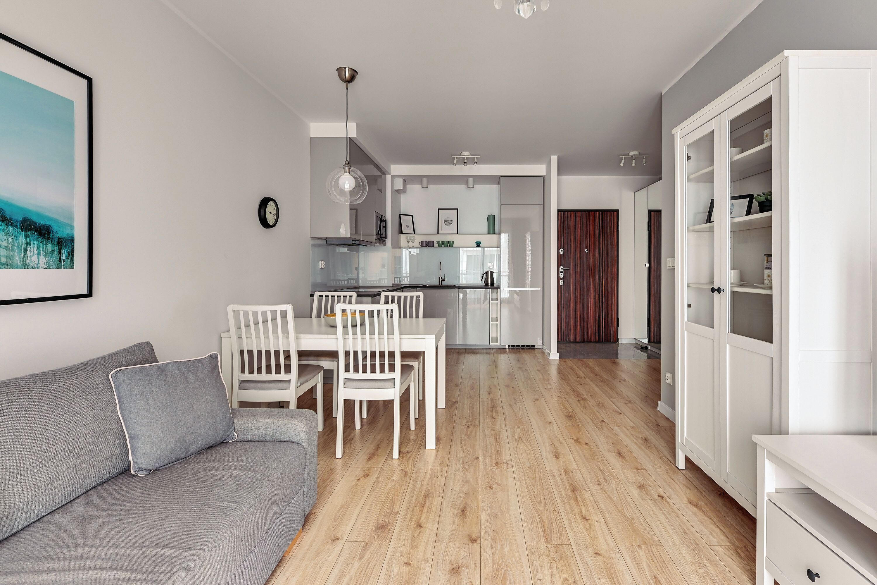 Un salon moderne avec un canapé gris, une table à manger avec des chaises, une cuisine en arrière-plan et un meuble blanc.