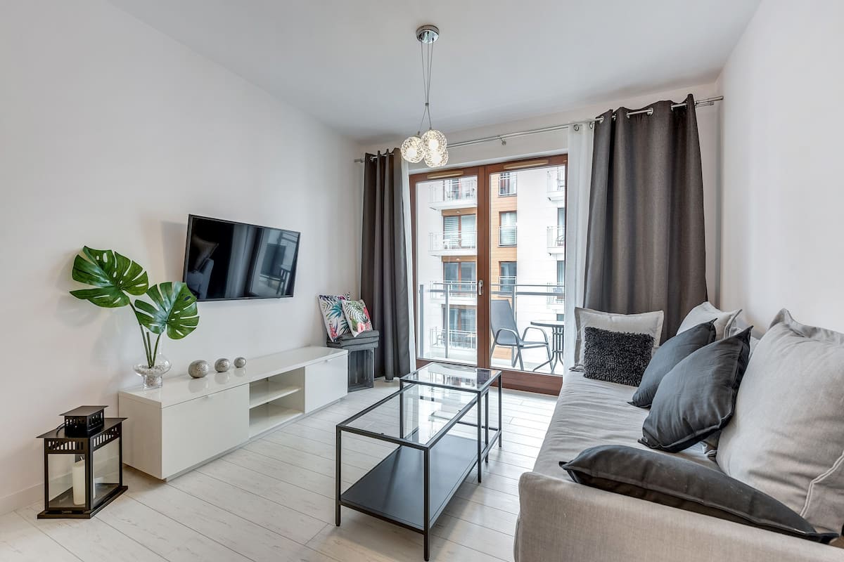 Een moderne woonkamer met een glazen salontafel, een witte tv-meubel, een grote plant en een bank met meerdere kussens.