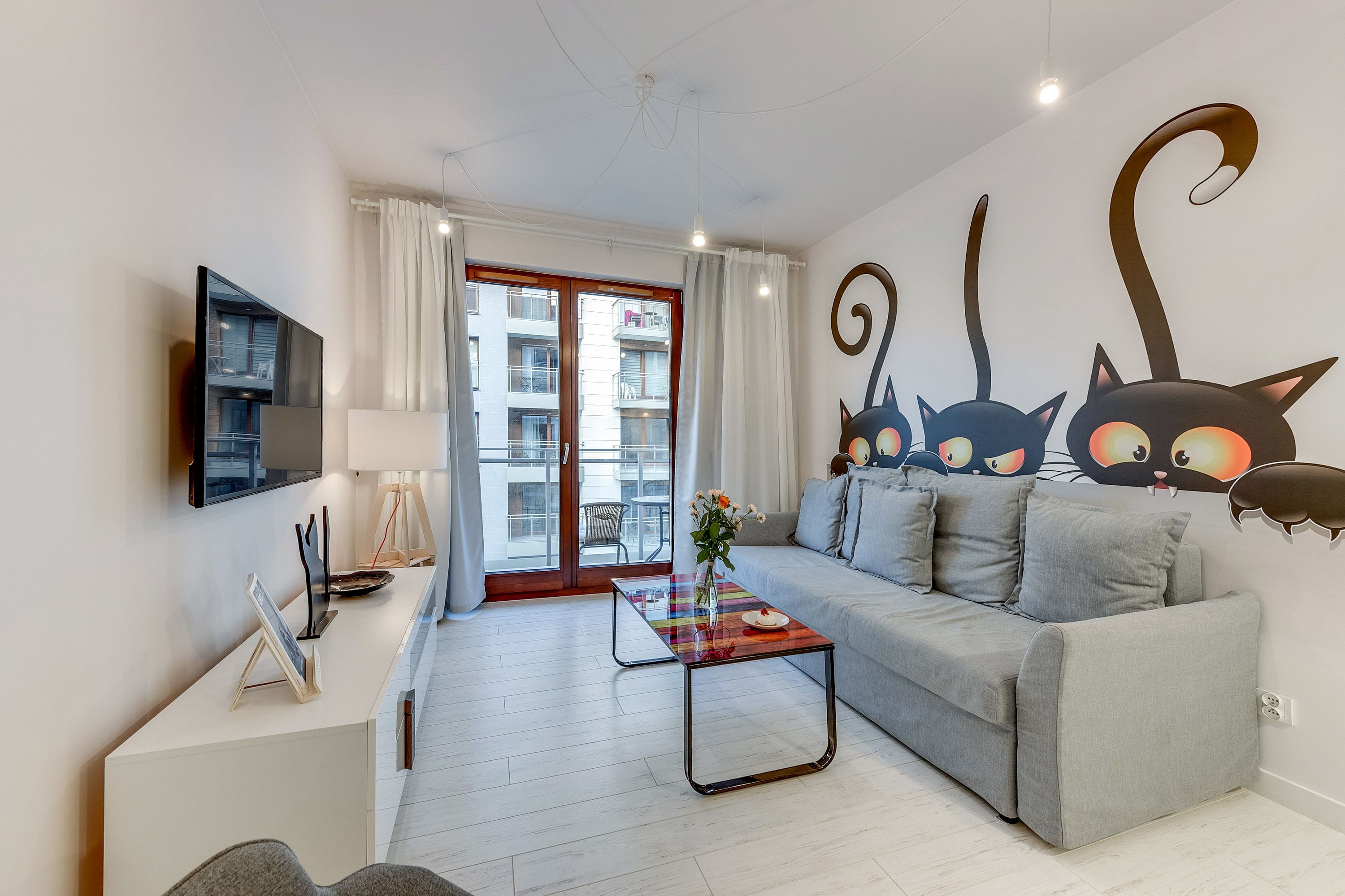 Un salon moderne avec un canapé gris, une table basse en verre, un téléviseur à écran plat et une grande fenêtre avec des rideaux.