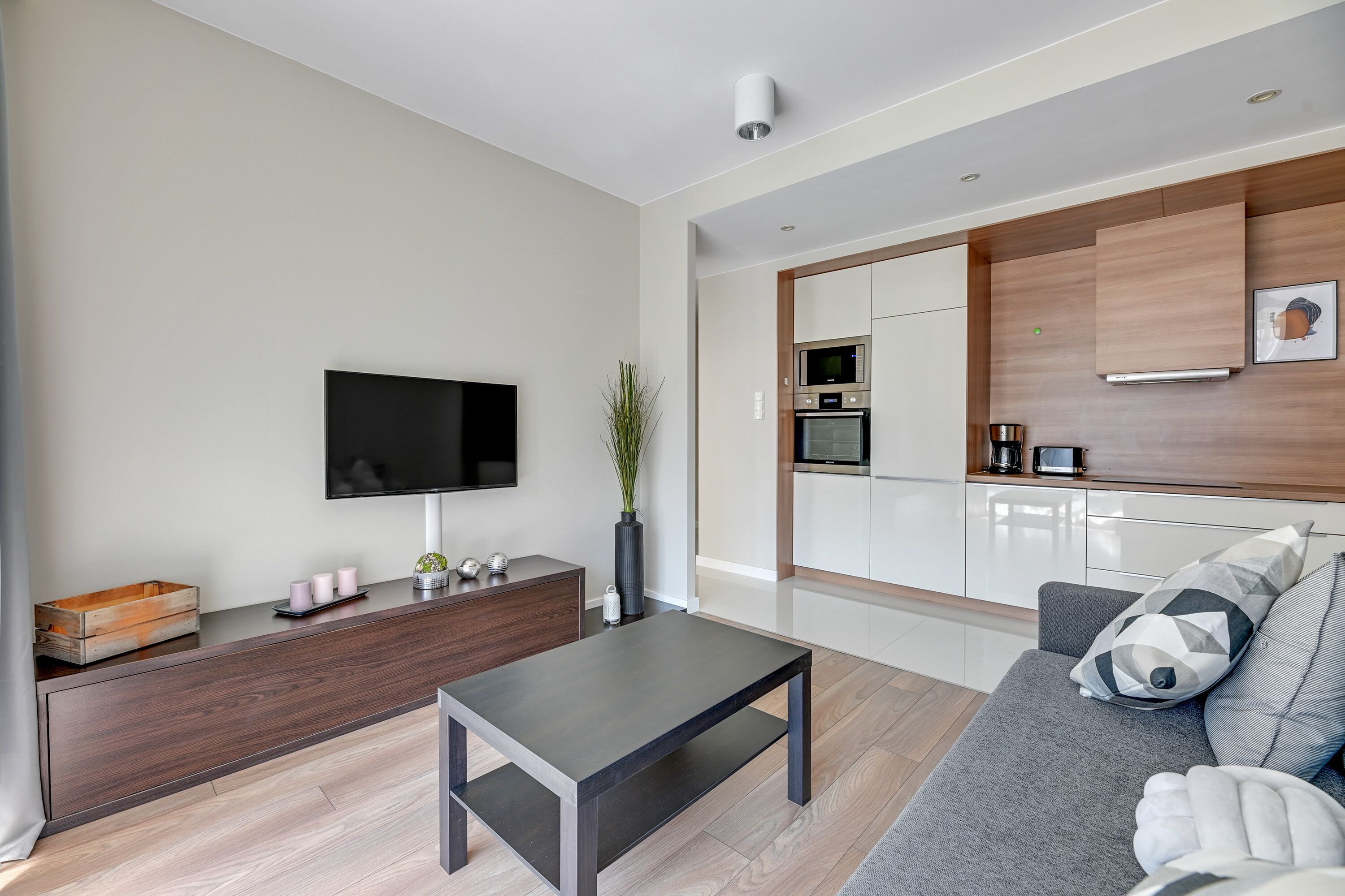 Een moderne woonkamer met een flatscreen televisie, een donker houten salontafel, een grijze bank met gekleurde kussens, en een keuken met witte kasten en een magnetron.