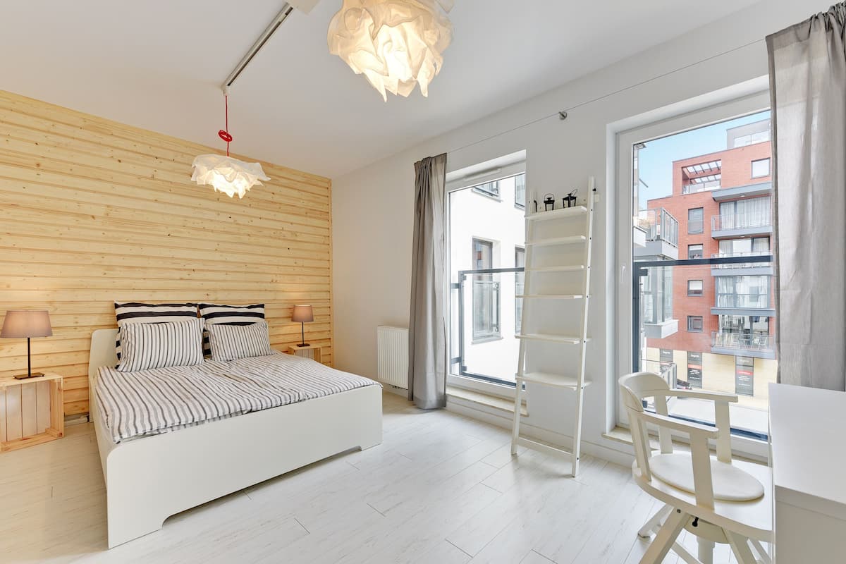 Een moderne slaapkamer met een bed, nachtkastjes, een stoel, een bureau, een raam met uitzicht op gebouwen en een bijzondere hanglamp.