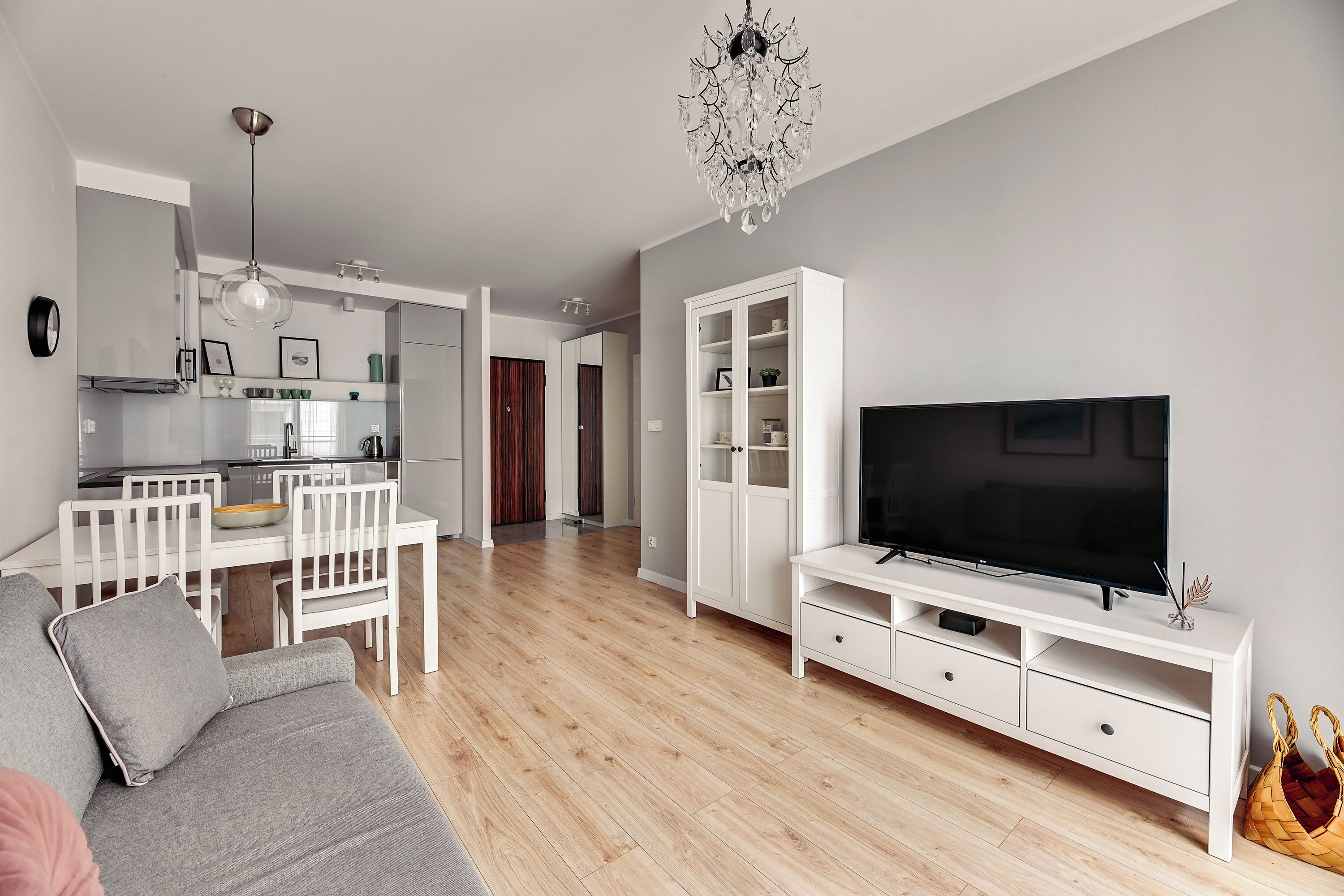 A modern living room with a grey sofa, a flat-screen TV on a white TV stand, a chandelier, and a kitchen in the background.