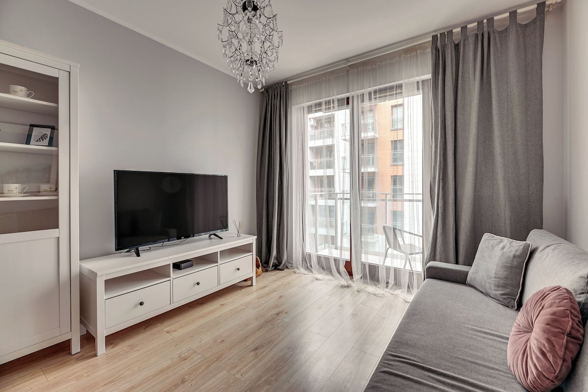 Een moderne woonkamer met een flatscreen televisie, een witte tv-kast, een grijze bank met kussens en een groot raam met grijze gordijnen.