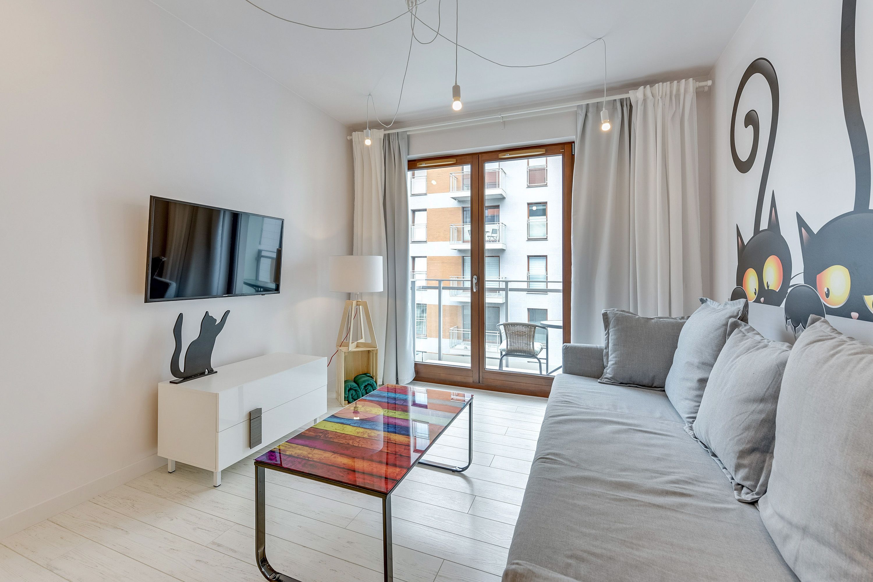 A modern living room with a sofa, a TV mounted on the wall, a colorful table, and a large window with curtains.