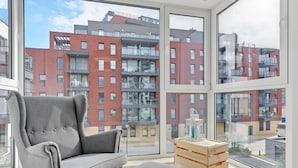 A modern living room with a grey armchair and ottoman, a wooden side table, and large windows offering a view of red brick apartment buildings. - NORDBYHUS Apartamenty Chmielna Park (Gdansk)
