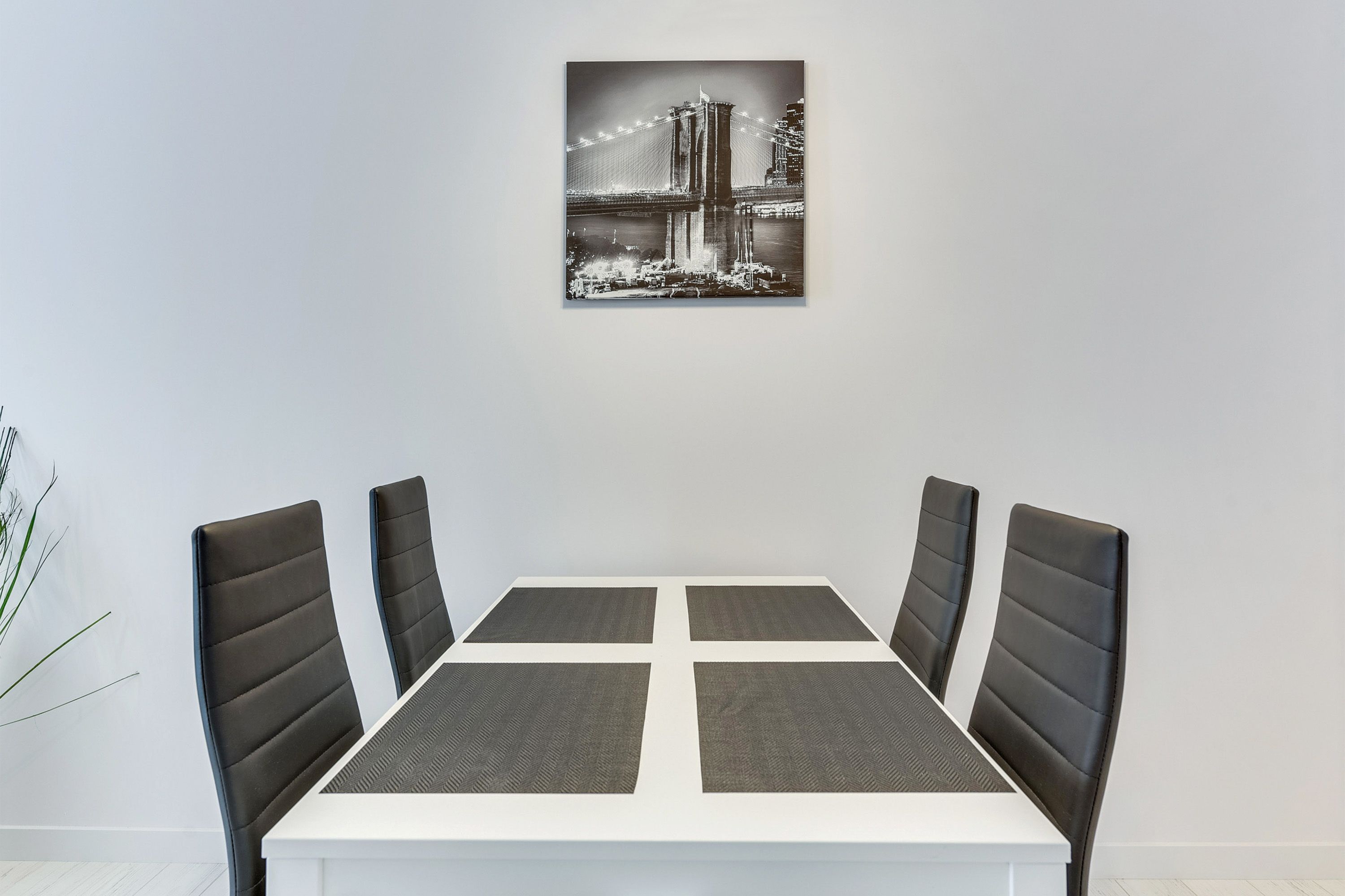 A dining area with a white table and four black chairs, a black and white photo of a bridge on the wall, and a plant to the left.