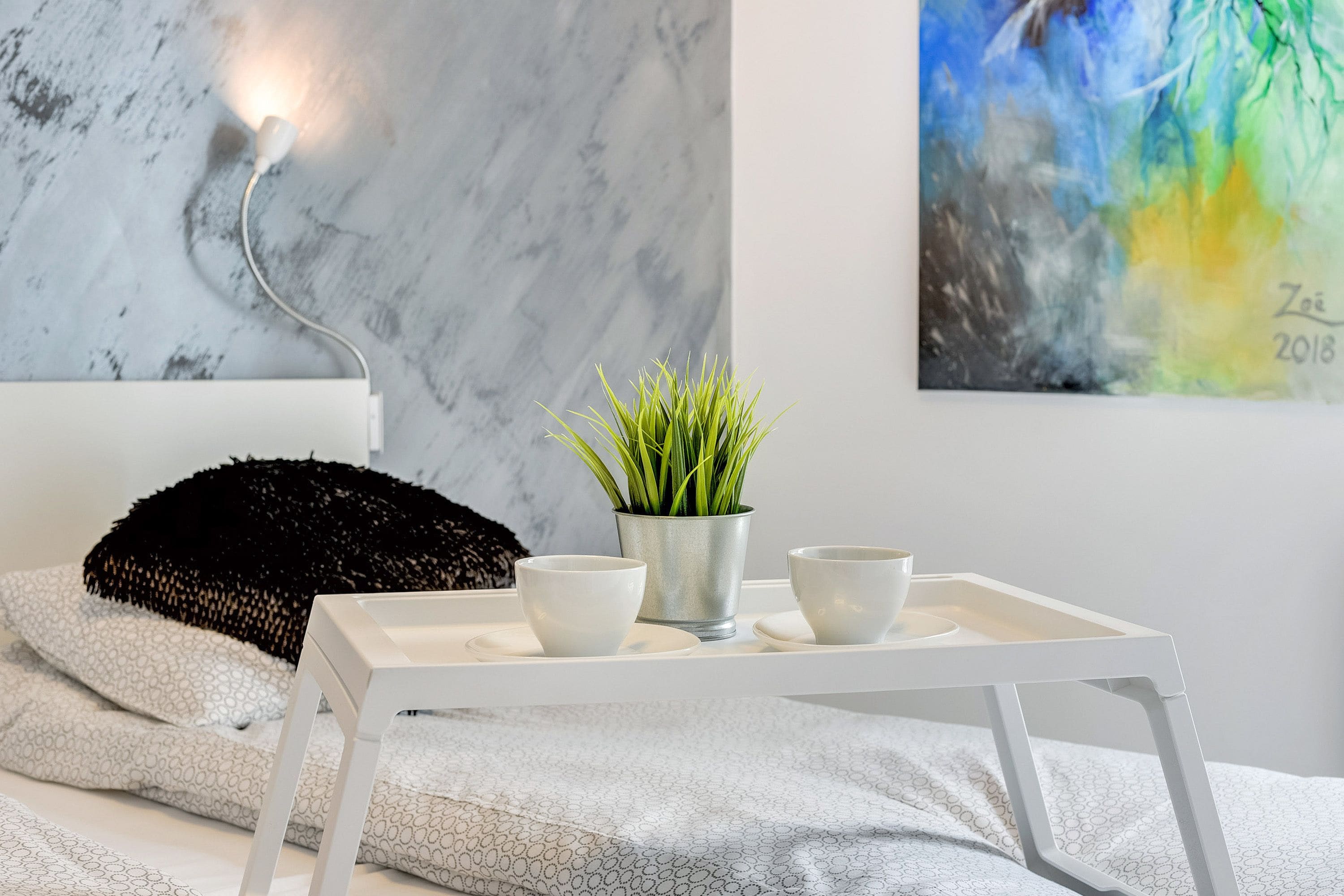 Ein ordentlich eingerichtetes Schlafzimmer mit einem weißen Bett, einem schwarzen, strukturierten Kissen, einer kleinen Topfpflanze und einem weißen Tablett mit zwei Schüsseln.