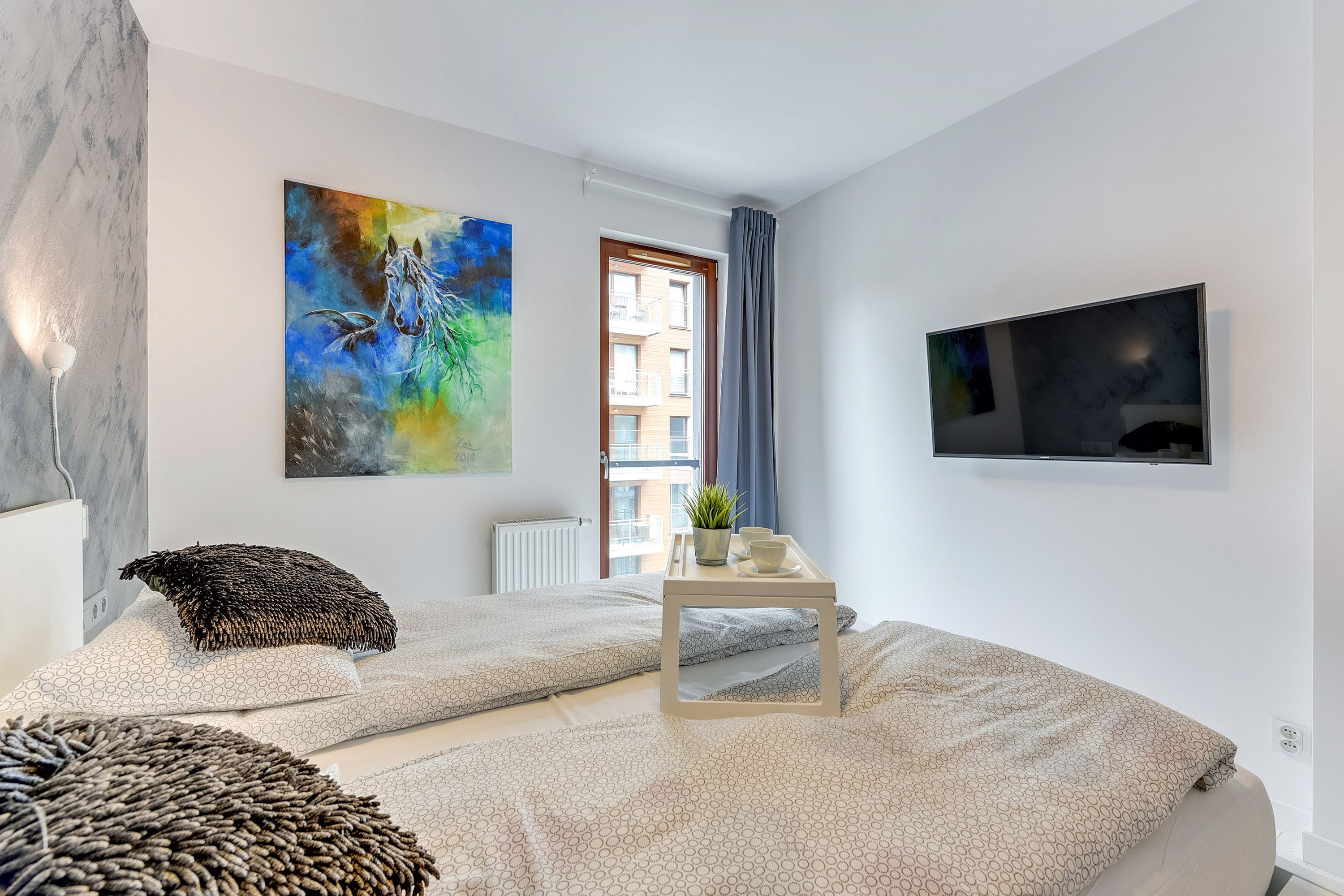 Un dormitorio moderno con cama, un cuadro, un televisor y una ventana con vistas a edificios.