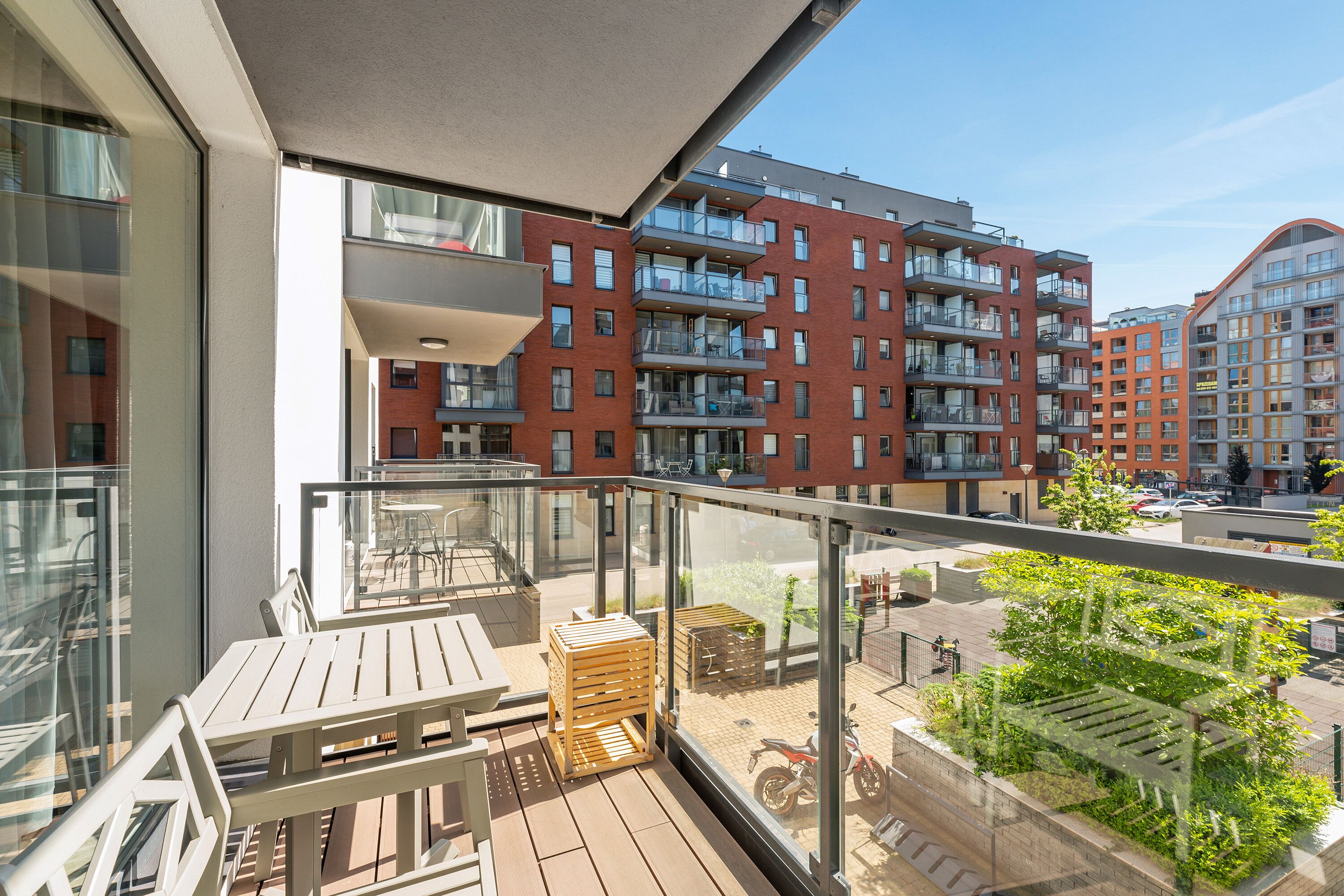 A balcony with a wooden table and chairs, a glass railing, and a view of red brick apartment buildings.
