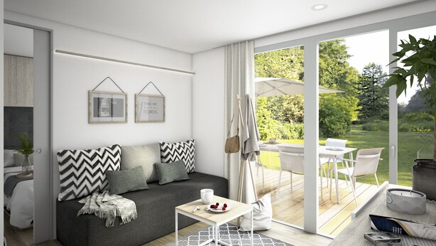 Ein modernes Wohnzimmer mit einem grauen Sofa, einem kleinen weißen Couchtisch und Blick auf einen Garten durch große Schiebeglasfenster.
