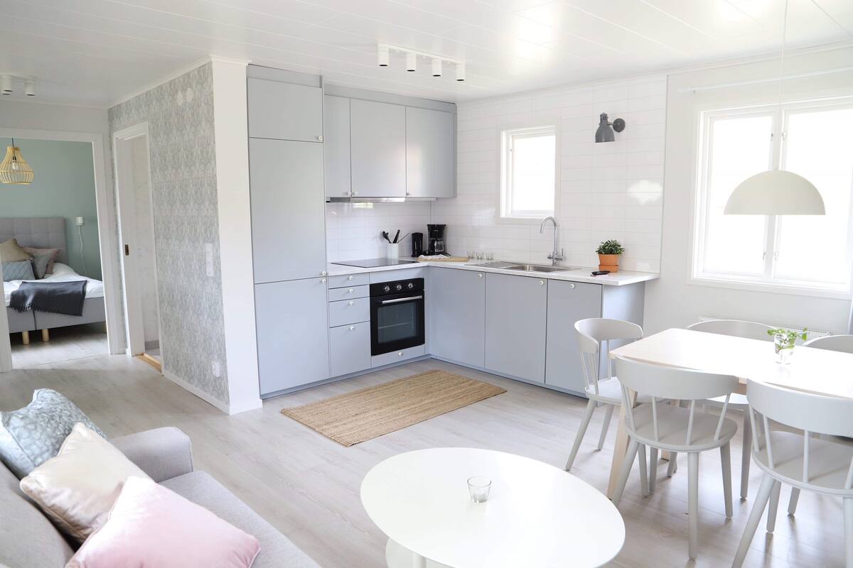 Cozinha moderna com armários brancos, sala de jantar com mesa e cadeiras brancas, e aconchegante sala de estar com sofá e almofadas.