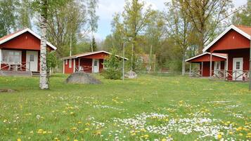 Tres cabañas de madera rojas con detalles blancos, ubicadas en un área cubierta de césped, diente de león y árboles de abedul.