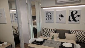 Ein moderner Essbereich mit Tisch für zwei Personen, einer Couch mit gemusterten Kissen und Wandbildern, darunter ein geometrisches Design und ein Motiv zum Thema Börse.