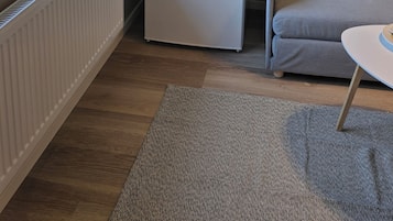 A compact living space with a white radiator, a small white appliance, a grey sofa, a white table, and a window with a view of houses and trees.