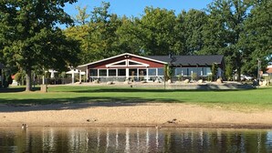 Ein Gebäude am Seeufer mit rotem Dach und großen Fenstern, umgeben von Bäumen und einem Sandstrand.