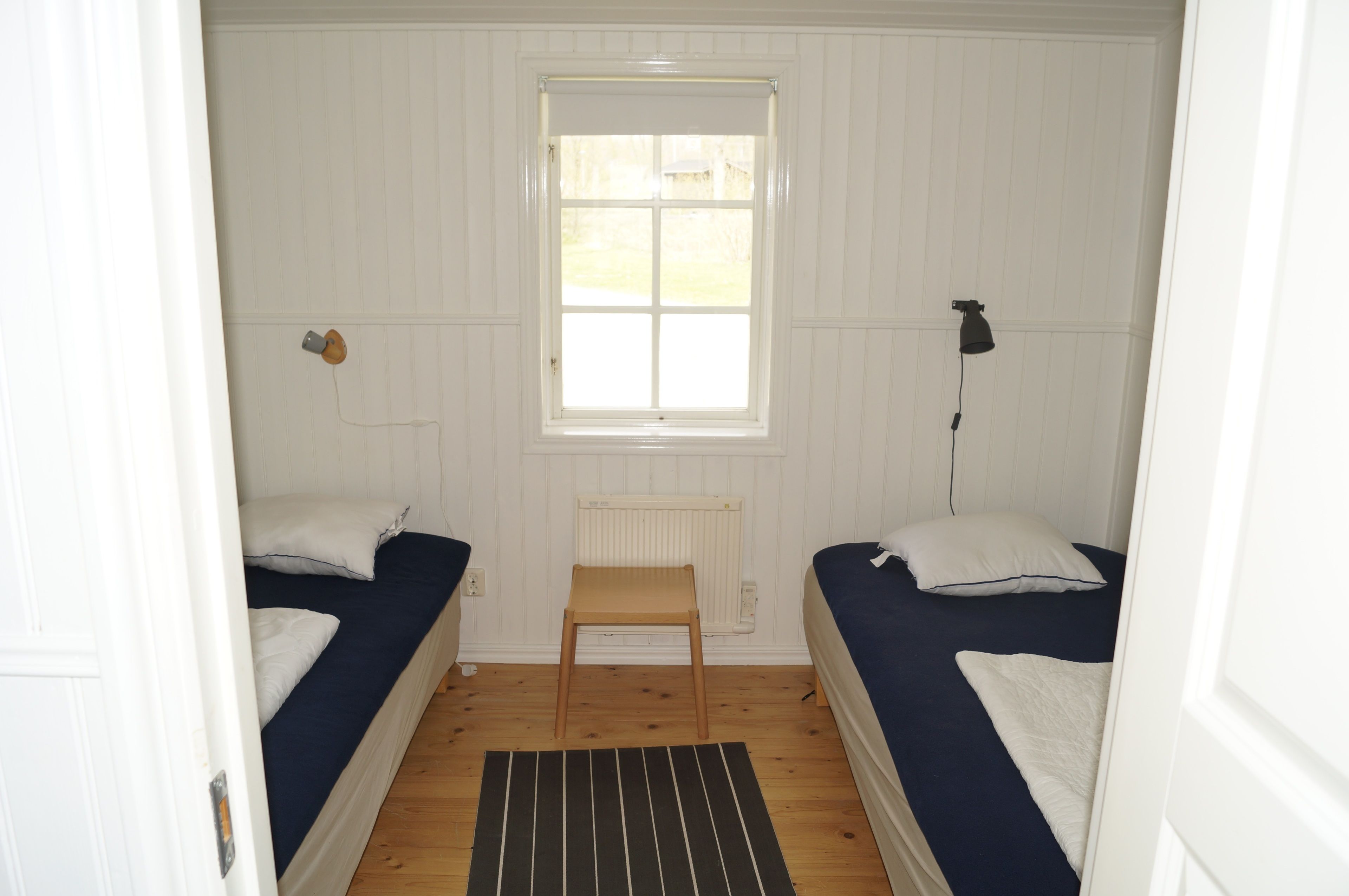 Un alojamiento pequeño y con una sola habitación, con dos camas individuales, una ventana, un radiador y un taburete de madera.