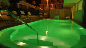 A swimming pool with a green light, surrounded by lounge chairs and a building in the background. - Residence Hotel Rubino (Riccione)