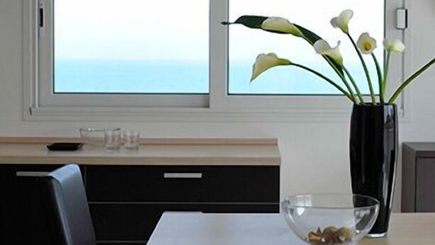 A dining area with a table, chairs, a vase with flowers, and a view of the sea through a window. - Residence Hotel Rubino (Riccione)