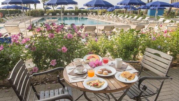 A poolside dining area with a table set for breakfast, overlooking a swimming pool with lounge chairs and umbrellas. - Fantinello Hotel (Caorle)