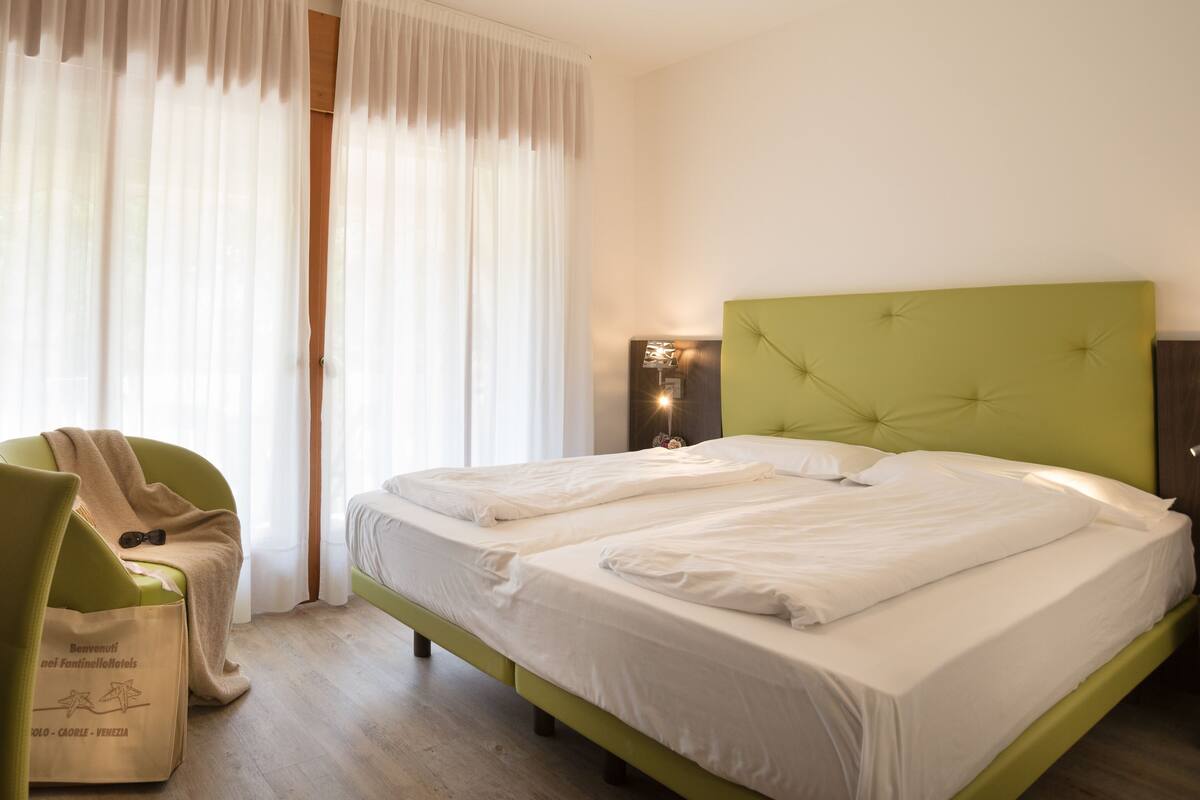 A hotel room with two beds, a green headboard, a bedside table, a chair, and a table with fruit.