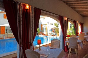 Un comedor con vista a la piscina, con una mesa para dos, sillas de mimbre y cortinas rojas.