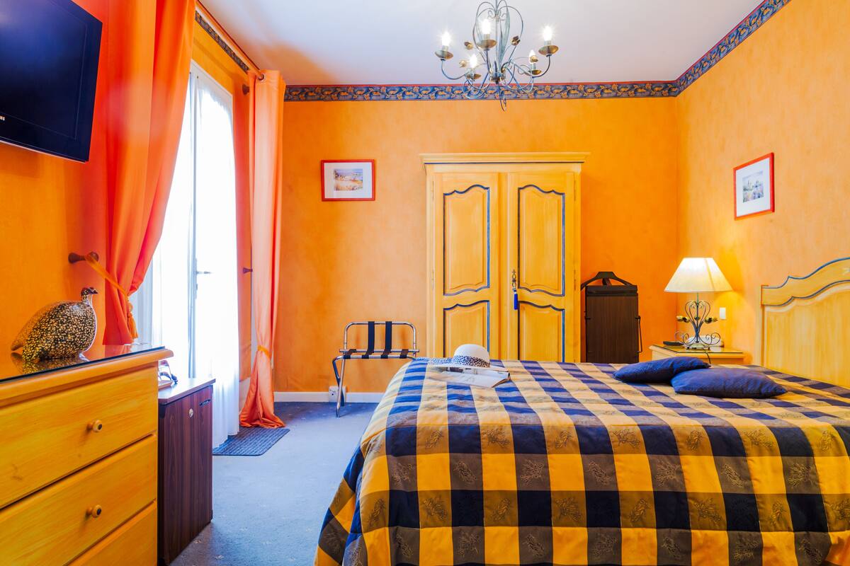 Quarto de hotel com colcha xadrez, cómoda de madeira, televisão e um candelabro.