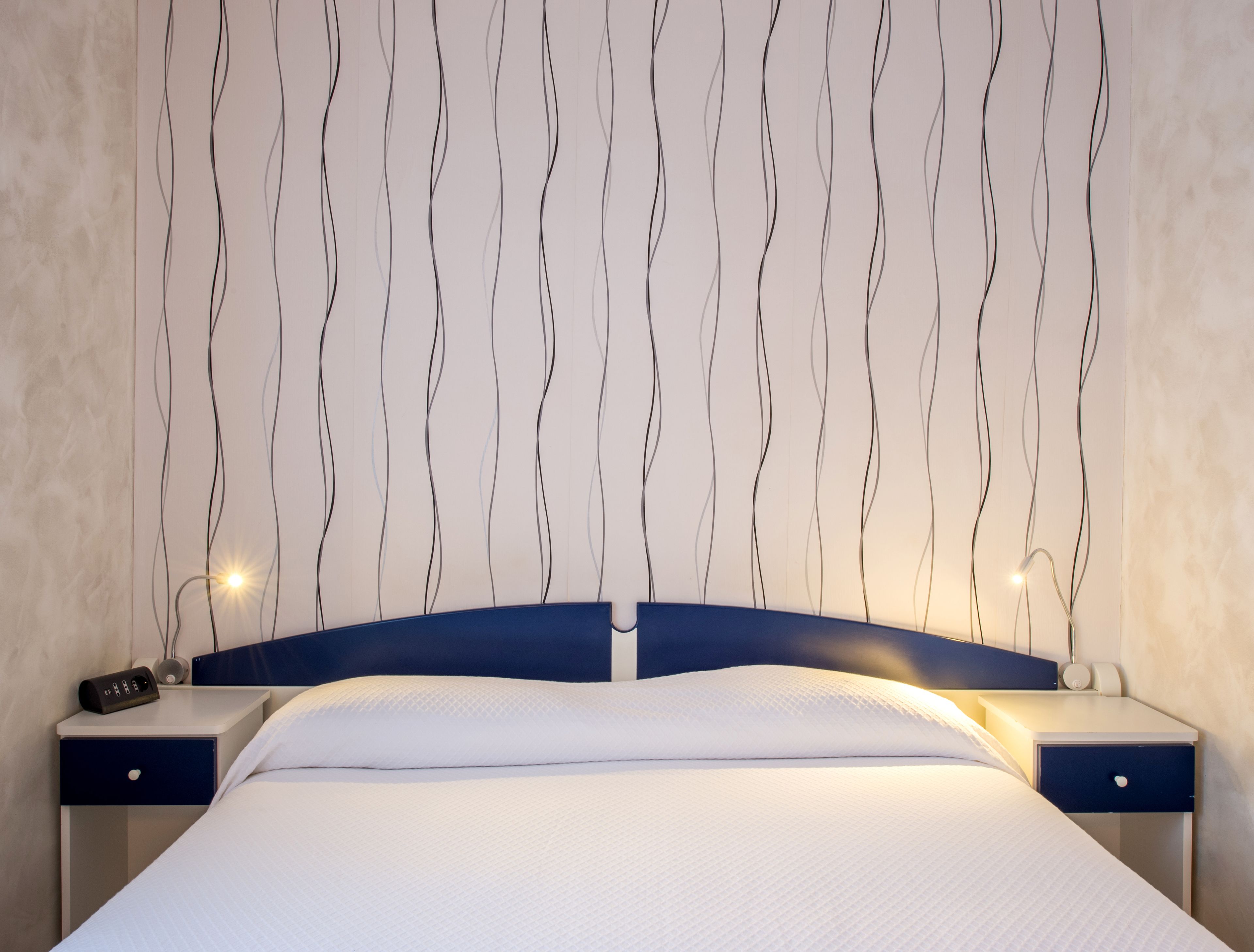 Een net opgemaakt bed met een wit laken en een blauw hoofdeinde. Er zijn twee nachtkastjes, elk met een lamp en een telefoon. De muur achter het bed heeft een opvallend golvend patroon.