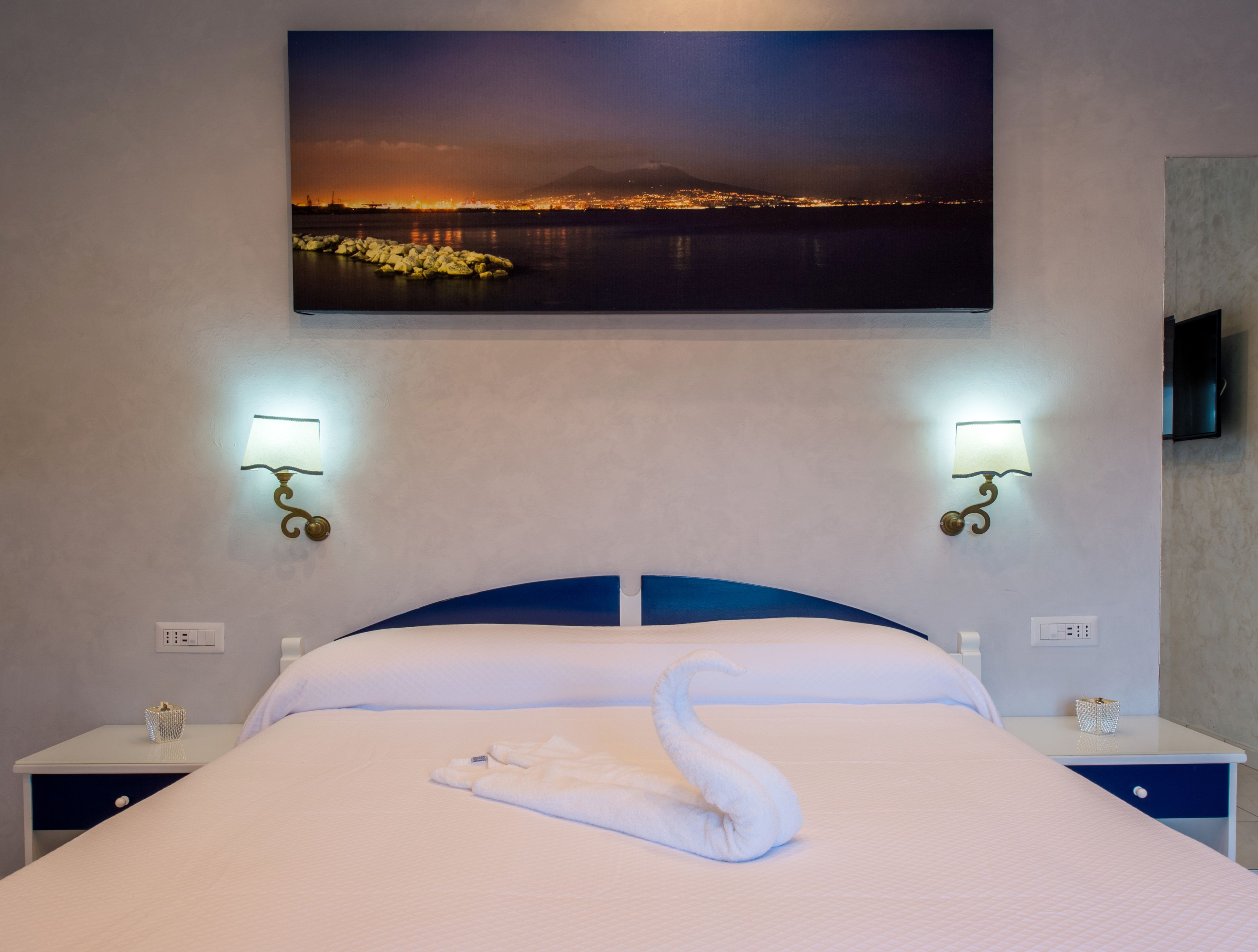 Ein Hotelzimmer mit einem Bett, zwei Nachttischlampen, einem Stadtbildgemälde und einem schwanenförmigen Handtuch auf dem Bett.
