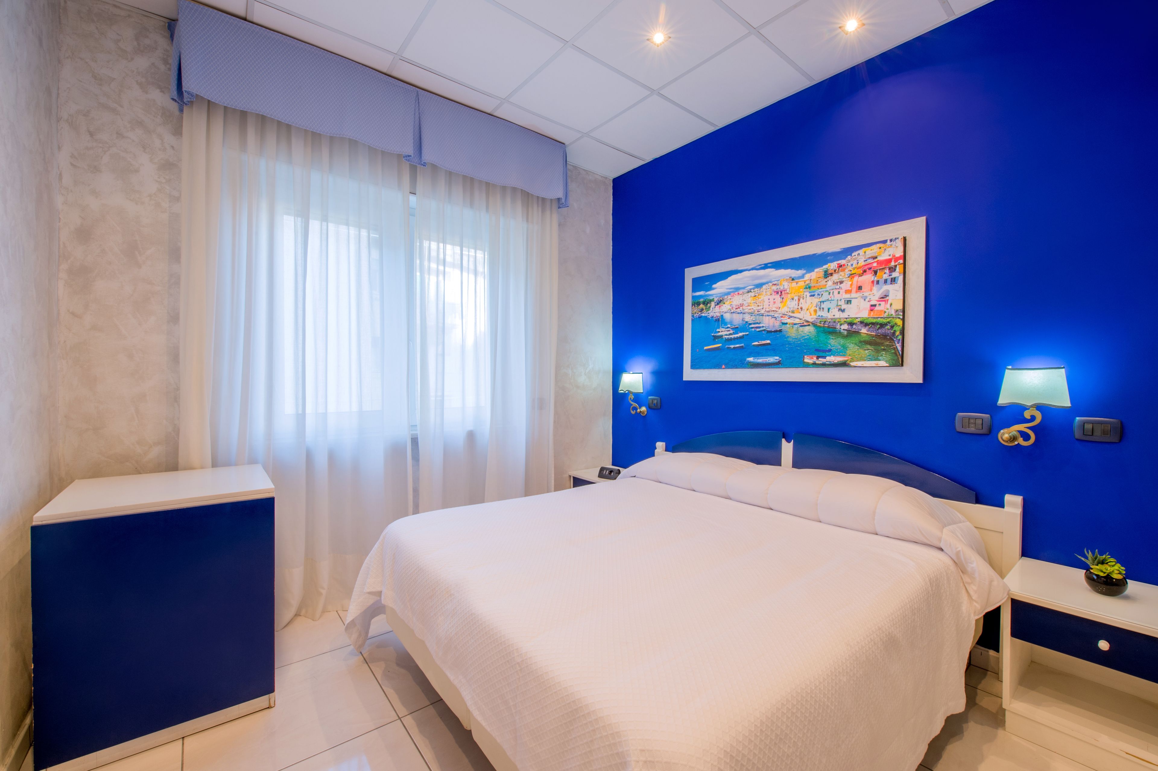 Hotelkamer met een groot bed, nachtkastje, een schilderij aan de muur en een raam met gordijnen.