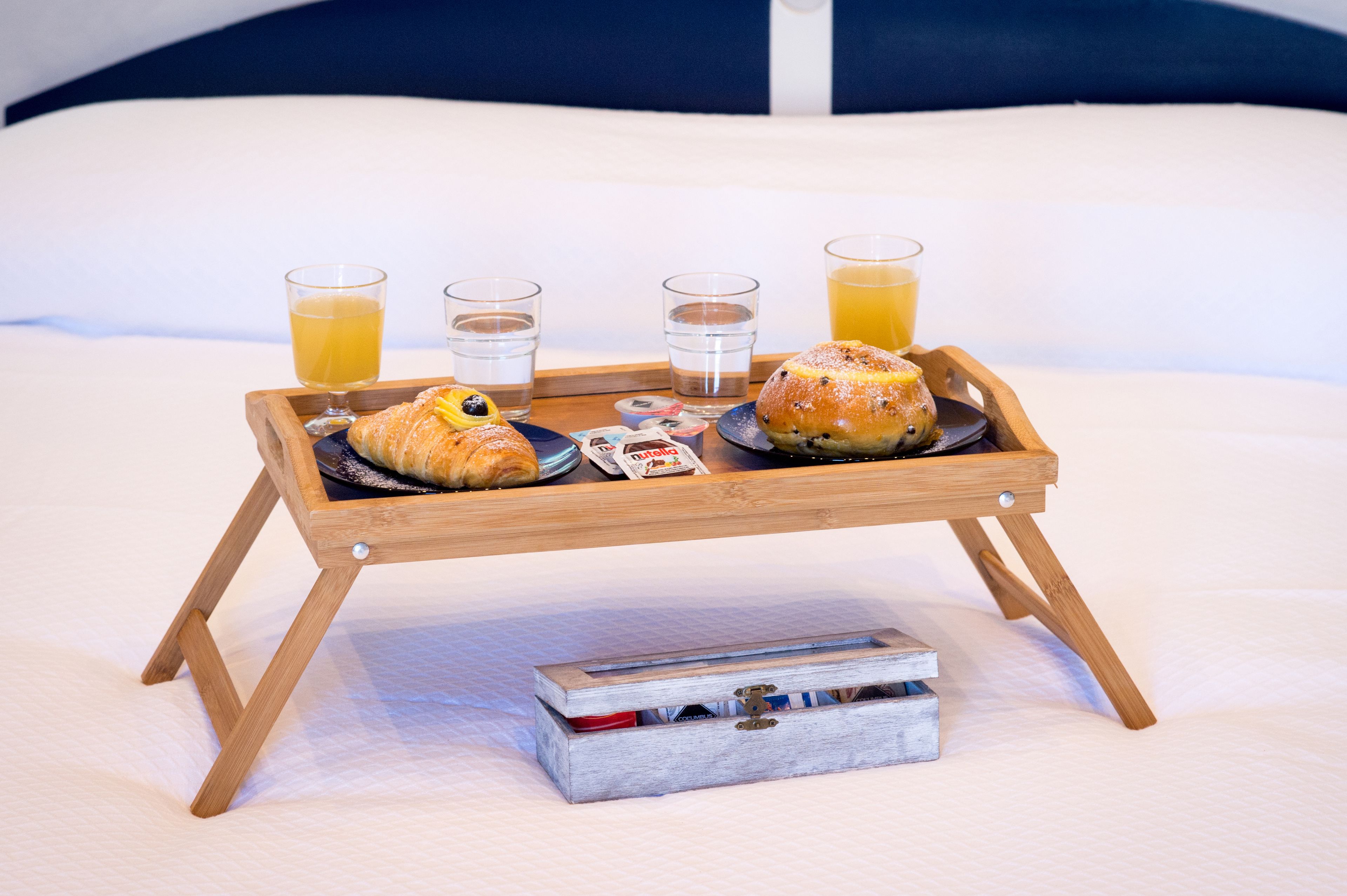 Un plateau de petit-déjeuner avec des croissants, du jus et des verres d’eau, posé sur un lit.