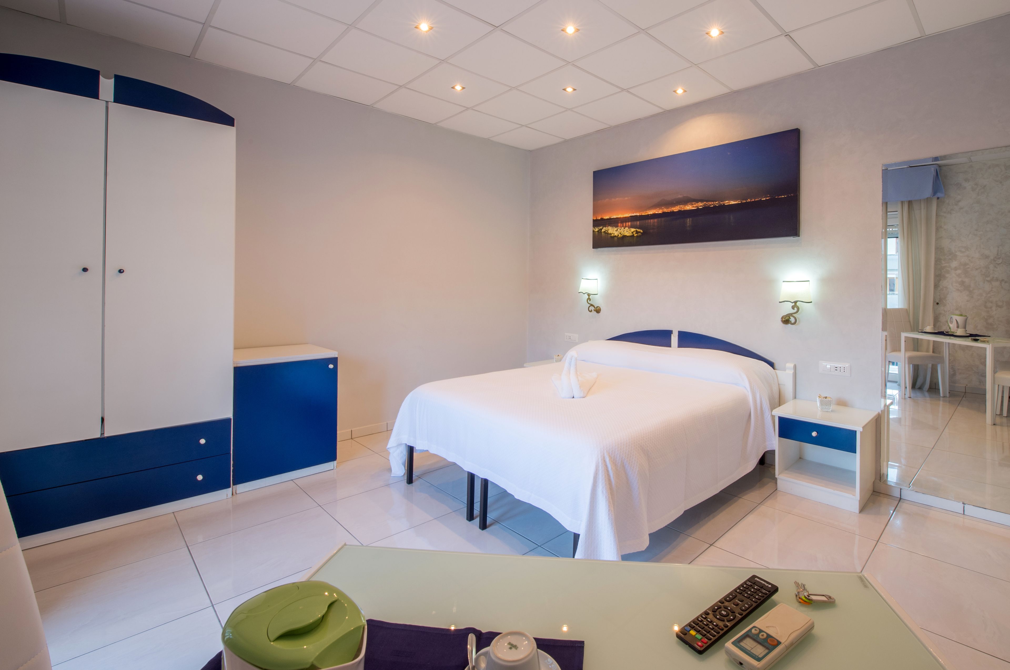A hotel room with a bed, bedside tables, a desk with a chair, a wardrobe, and a painting on the wall.