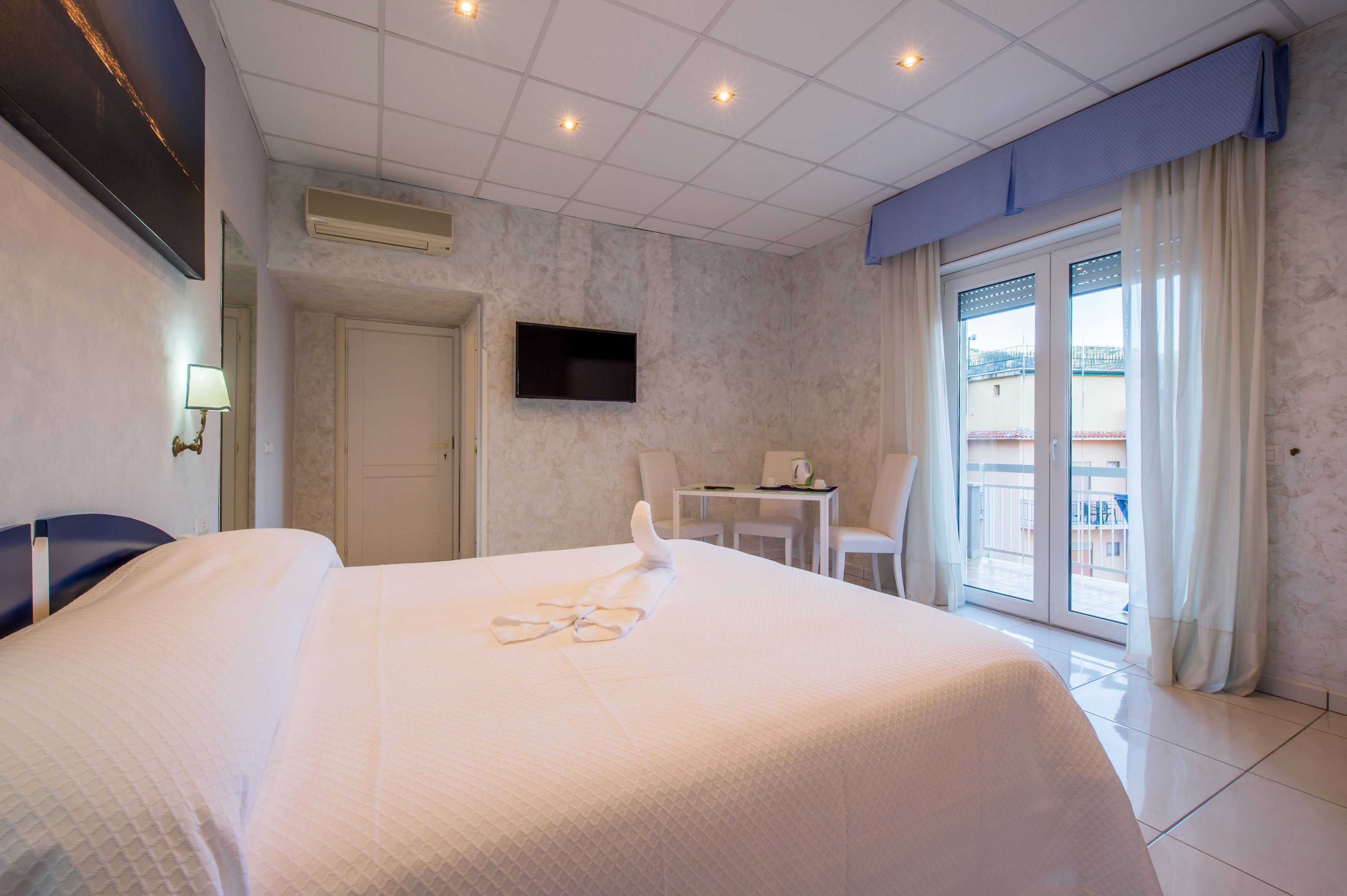 Habitación de hotel con una cama grande, un televisor de pantalla plana en la pared, una pequeña mesa con sillas y un balcón con cortinas.