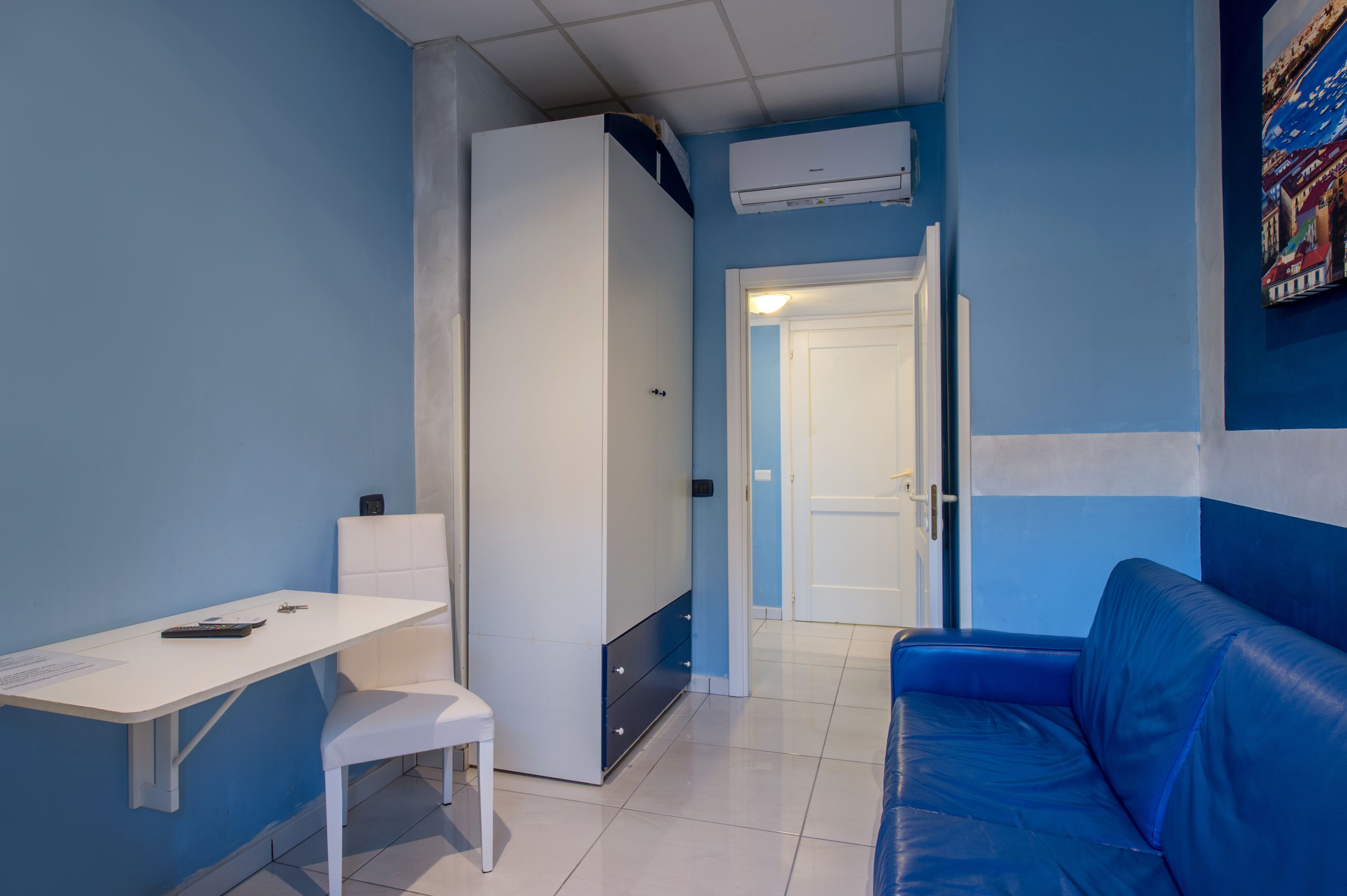 Een kamer met een wit bureau, een blauwe bank, een garderobe en een deur naar een andere kamer.
