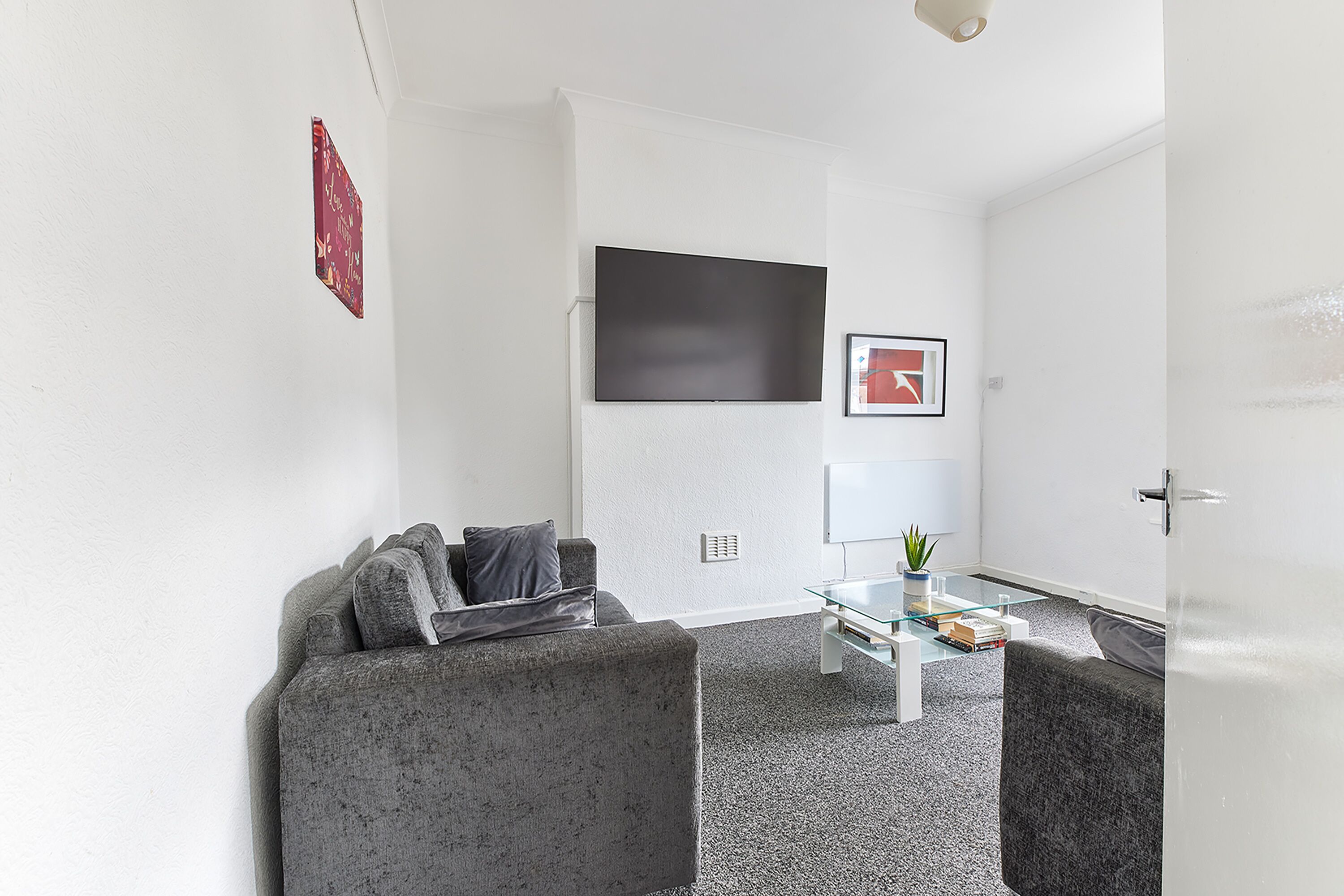 A modern living room with a grey sofa, a glass coffee table, and a flat-screen TV mounted on the wall.