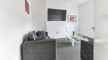 A modern living room with a grey sofa, a glass coffee table, and a flat-screen TV mounted on the wall.
