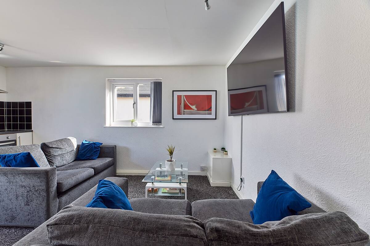 A modern living room with grey sofas, a glass coffee table, and blue cushions. There is a window with blinds, a framed picture on the wall, and a white side table with a plant.