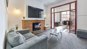 Una sala de estar con un televisor de pantalla plana sobre una chimenea de ladrillo, sofás grises, una mesa de centro de vidrio y un amplio ventanal corredizo que da a un espacio exterior.