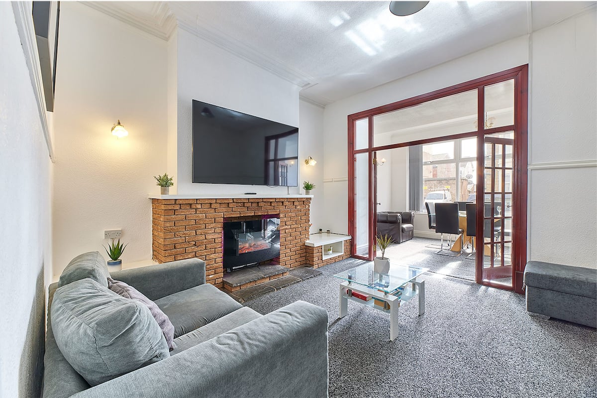 A living room with a flat-screen TV mounted above a brick fireplace, grey sofas, a glass coffee table, and a sliding glass door leading to an outdoor area.